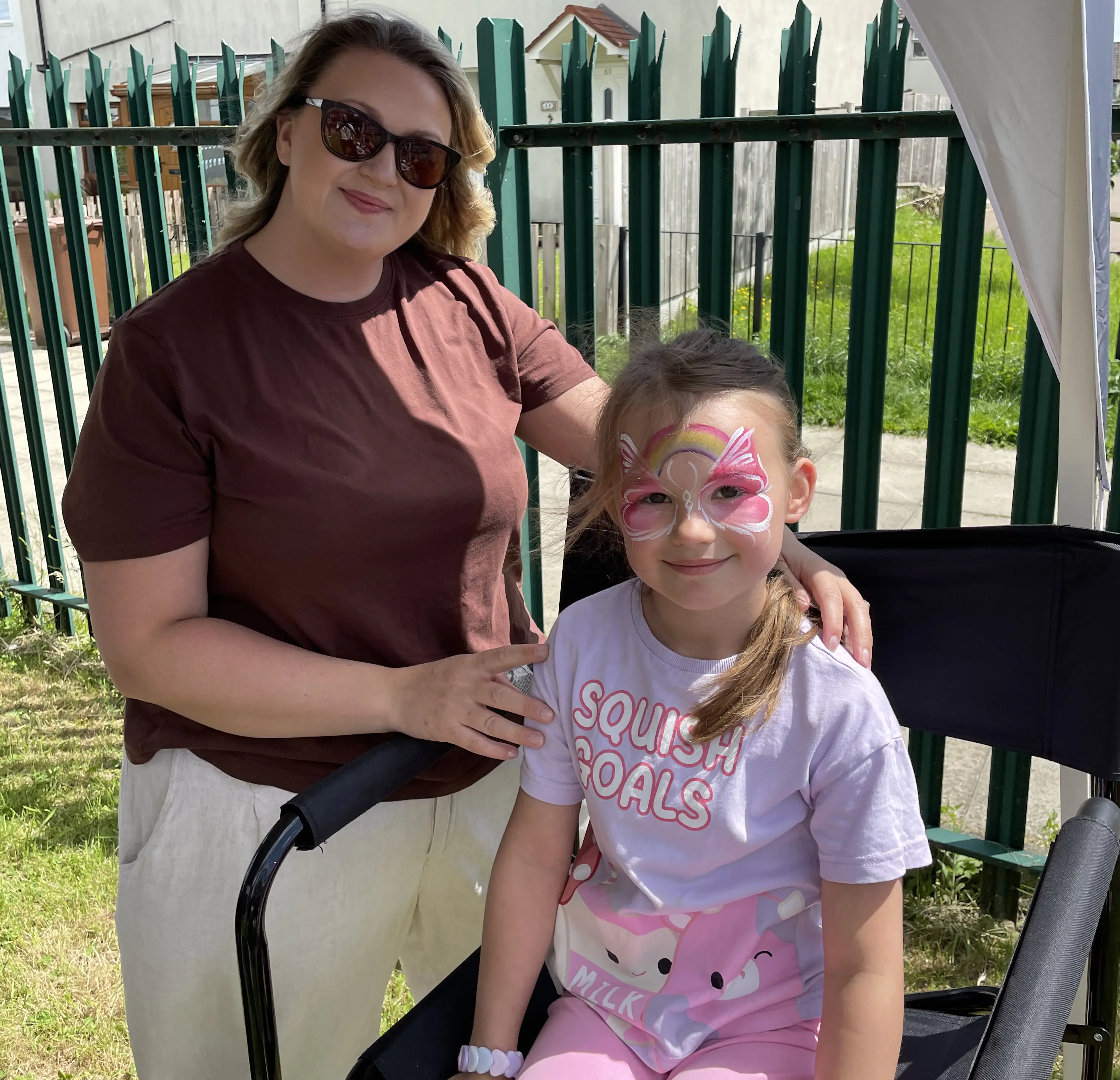 Blonde woman wearing sunglasses stands with her daughter who has had her face painted with a pink butterfly at Community Shop Swarcliffe.