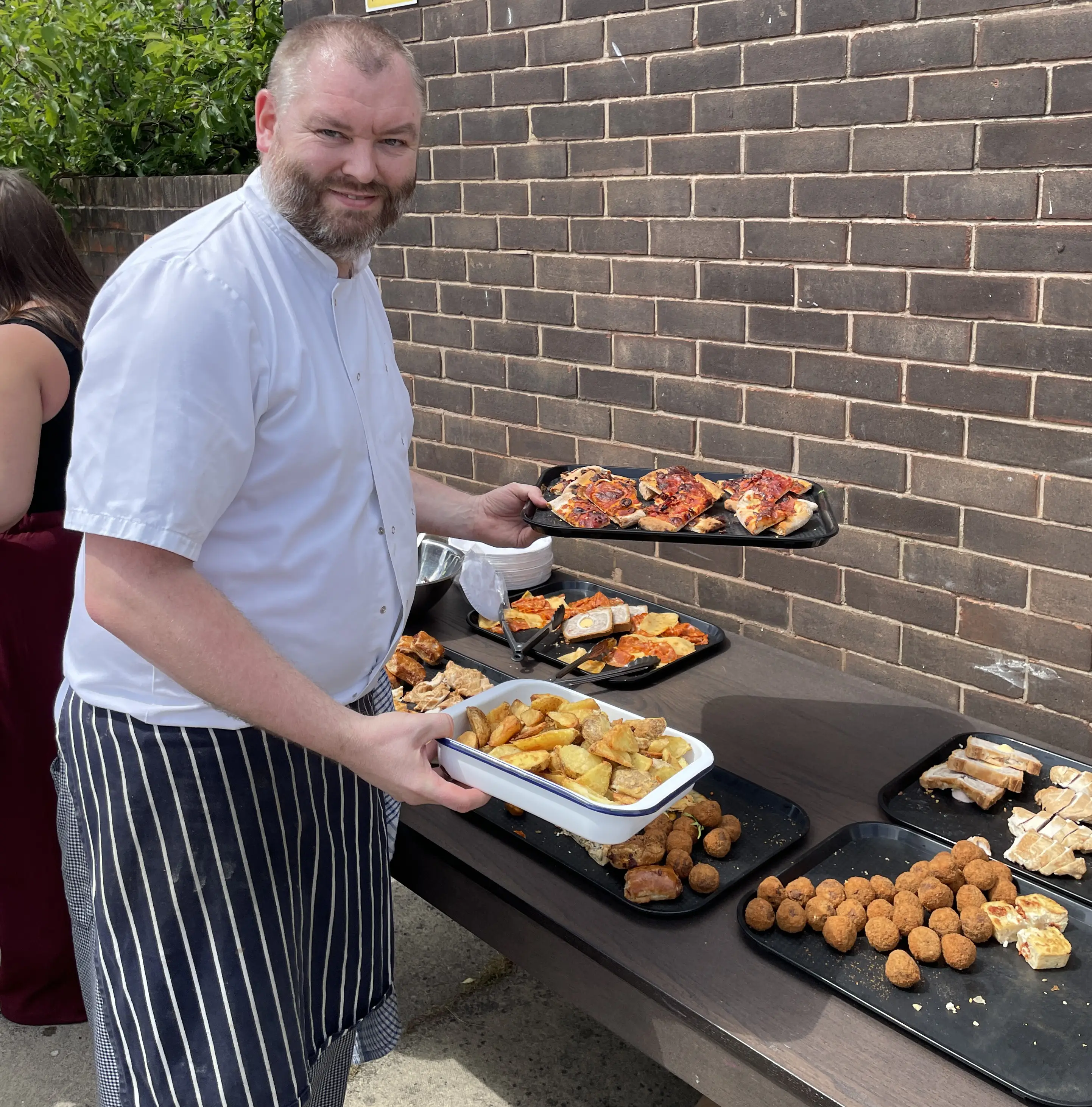 A chef in uniform places plates of potato wedges and pizza onto a table with scotch eggs and sandwiches.