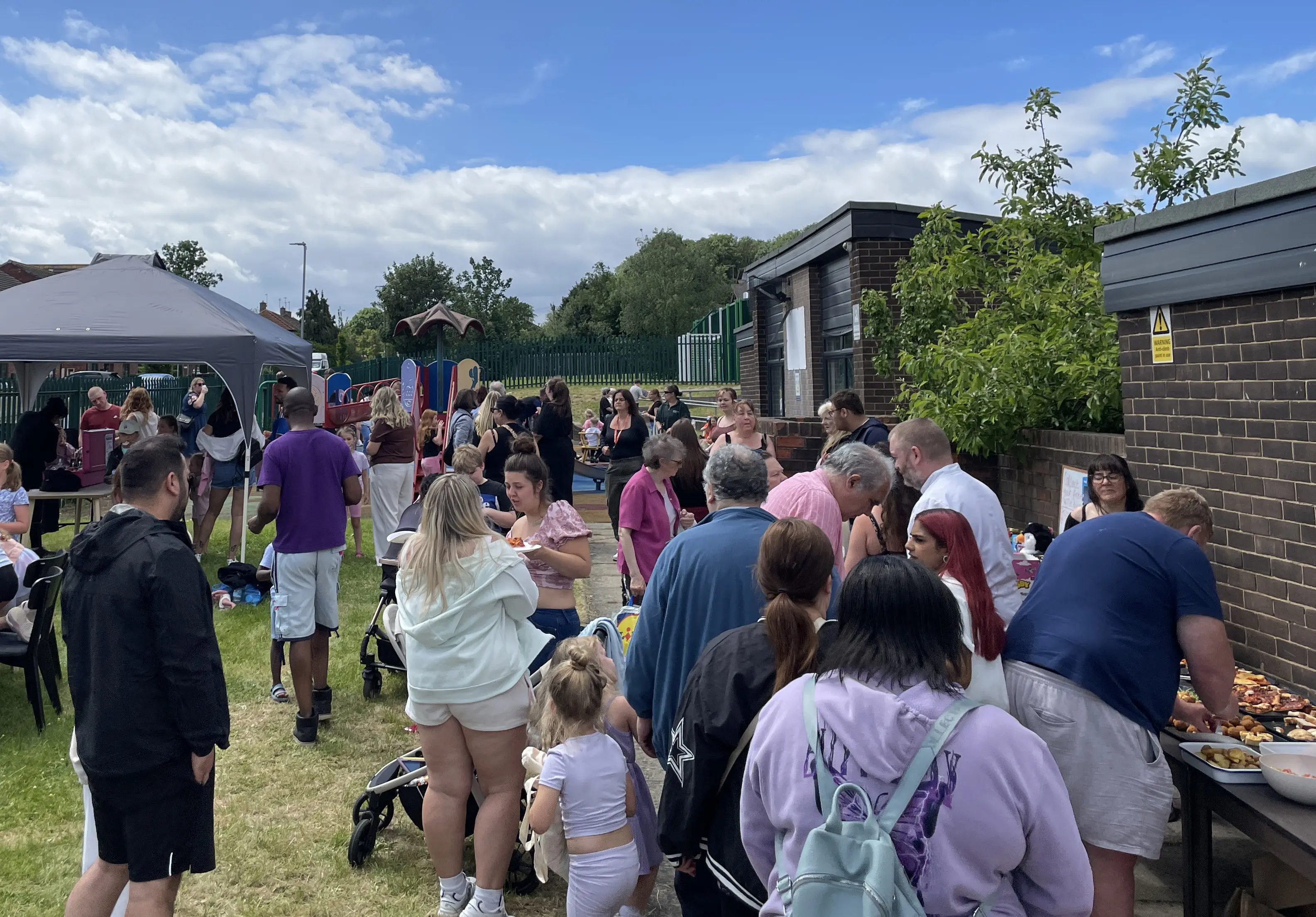Dozens of people in summer clothes attending Community Shop Swarcliffe's 1st Birthday party with party food, face painting and a children's playground.