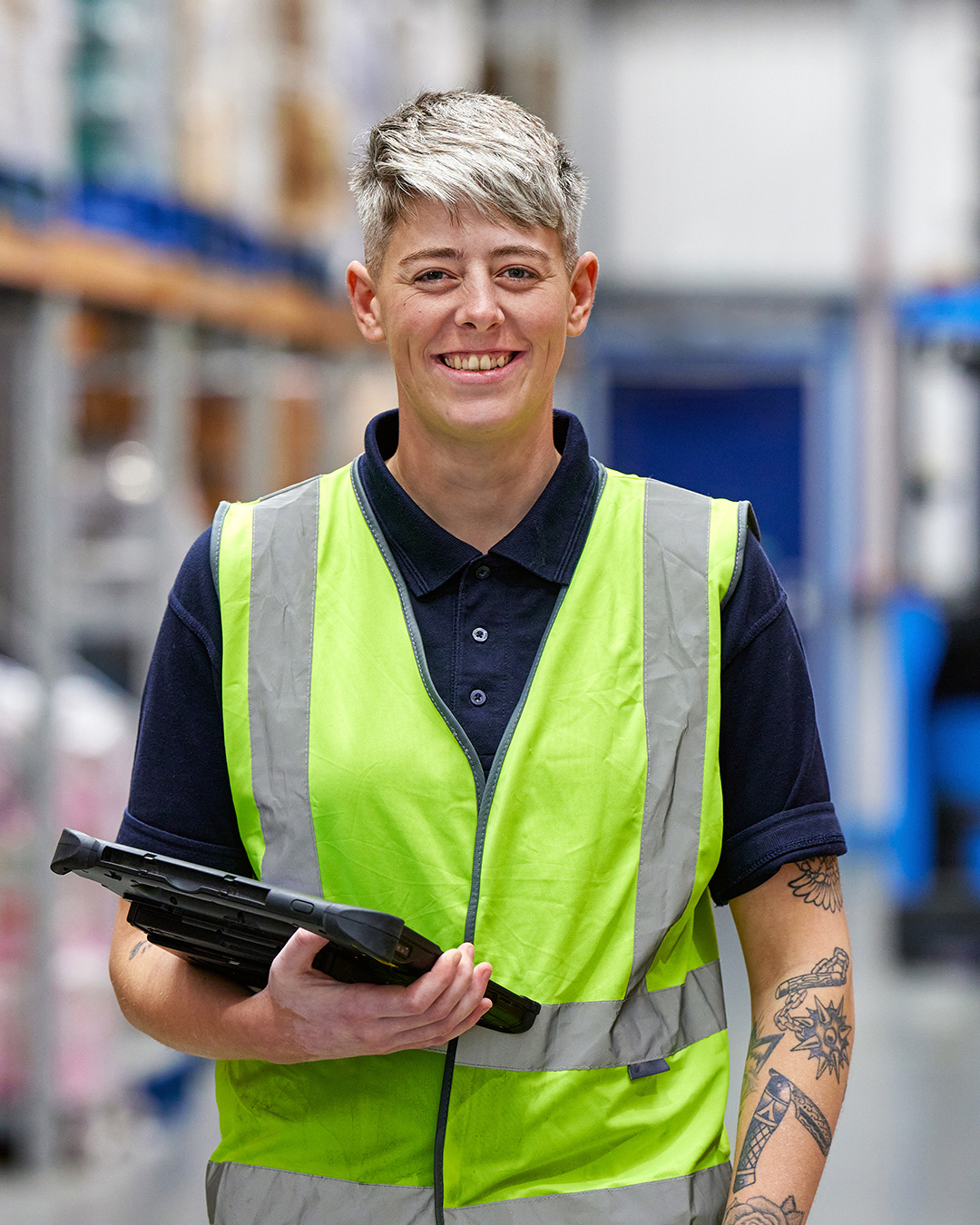 Company Shop Group warehouse operative holds a clipboard and smiles.