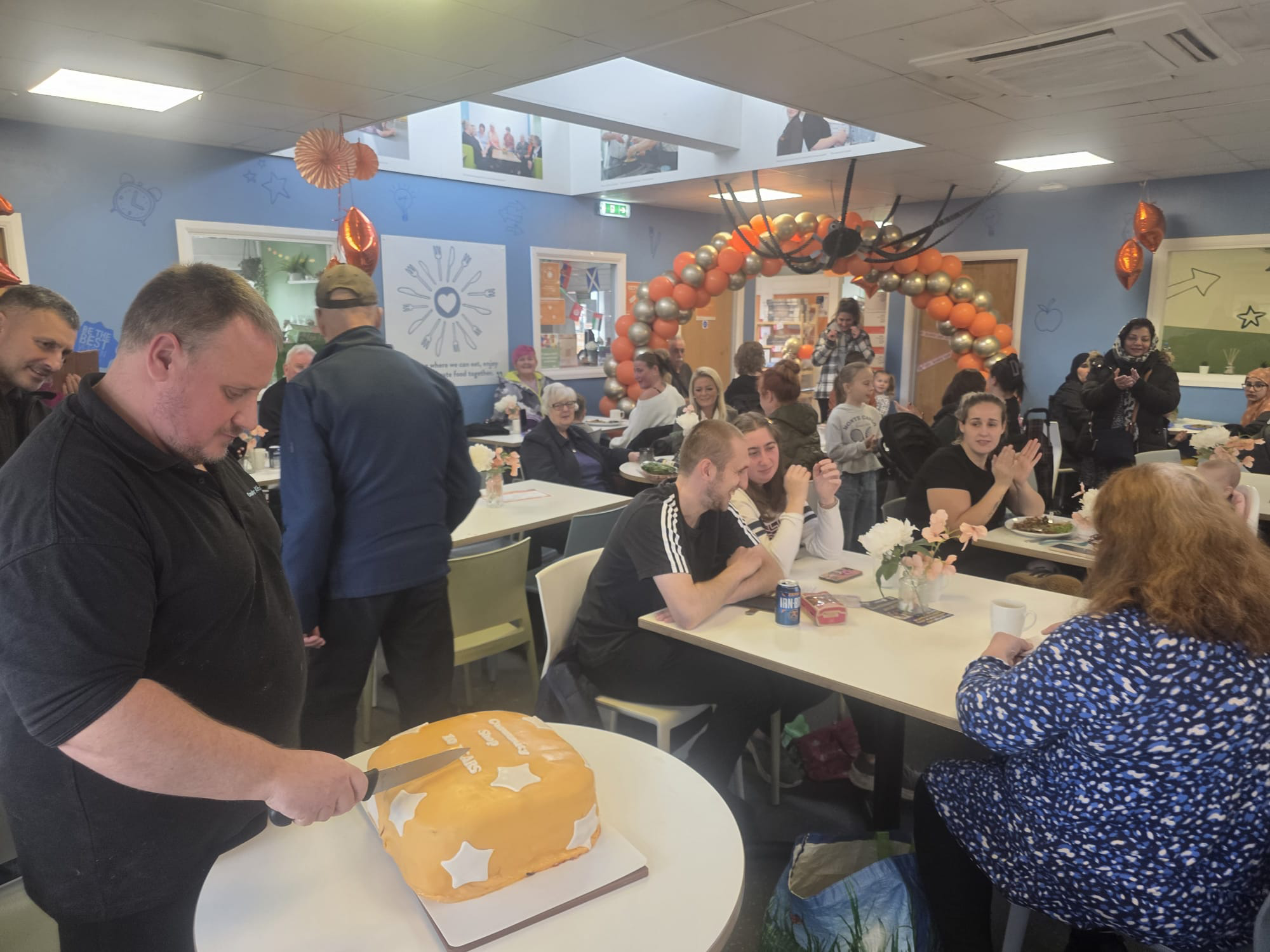 A group of people at Community Shop Athersley cheer as a chef cuts an orange birthday cake.