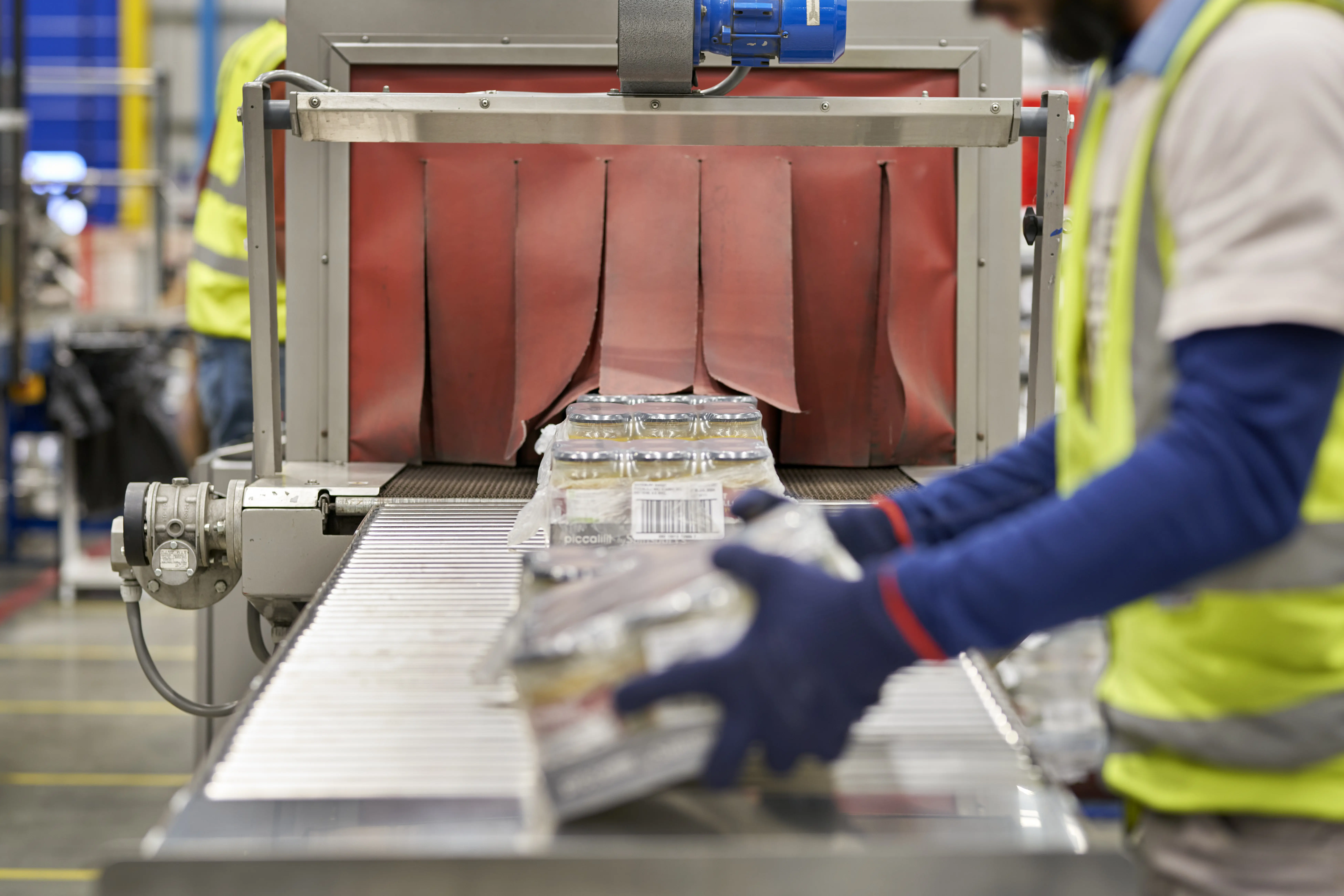 Jars of sauce wrapped in plastic are being placed onto a metal conveyor belt and passing through red conveyor skirting.