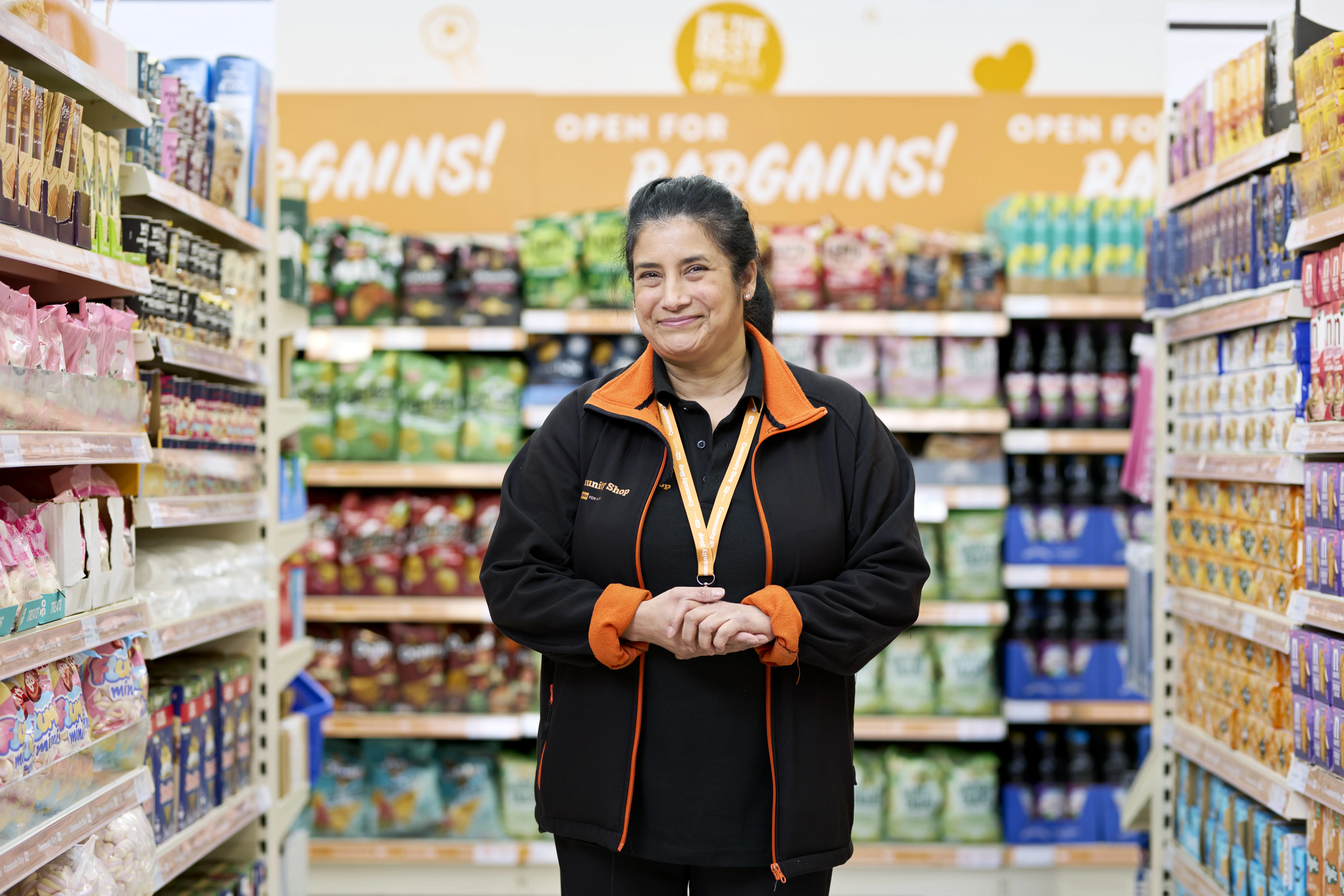 Smiling woman wearing a black and orange Community Shop fleece standing in the middle of a shop aisle stacked with cereal, tins of beans and toiletries.