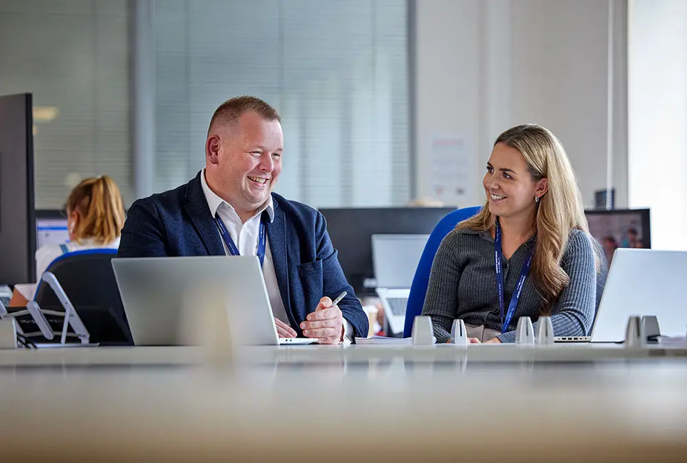 Male and female Company Shop Group colleagues sat at desks smiling as they collaborate on work.