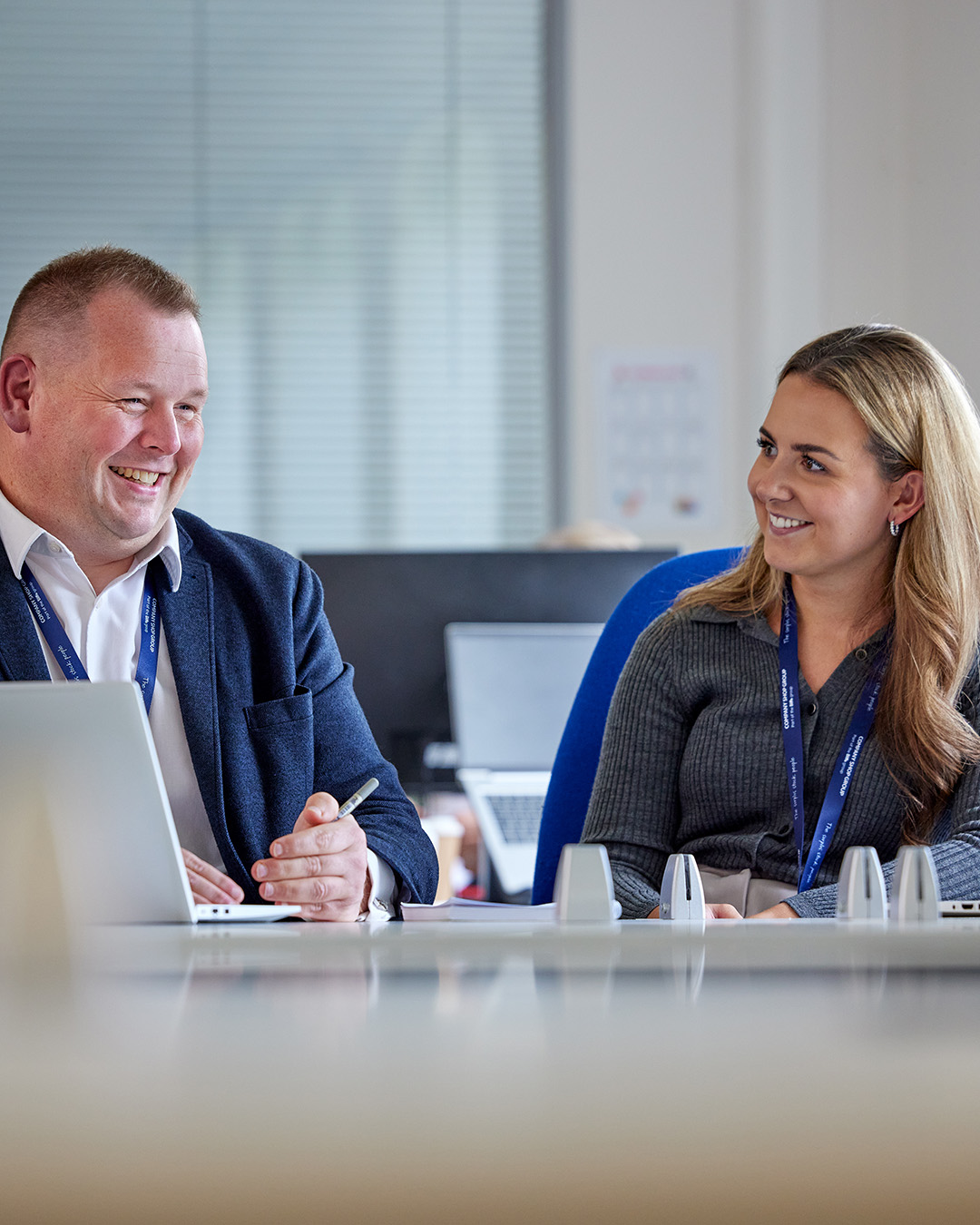 Male and female Company Shop Group colleagues sit at desks smiling as they collaborate on work.