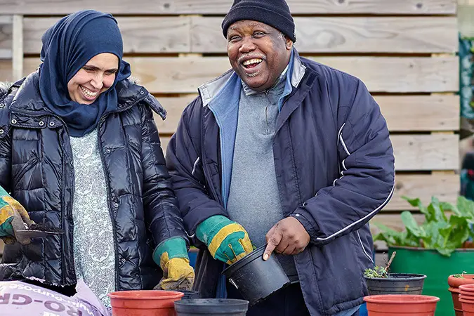 A woman wearing a dark blue hijab and shiny black puffer jacket laughs with a man wearing a woolly hat as they plant seeds at an allotment.