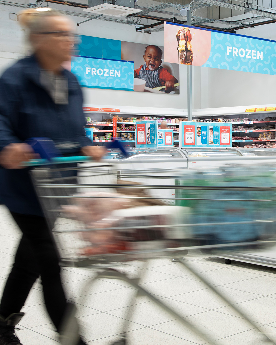 A woman pushes a trolley full of food through the frozen section of a Company Shop.