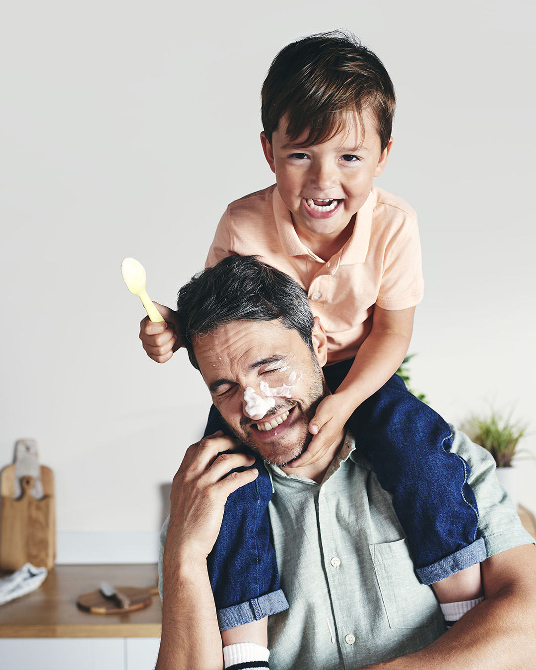 A young boy laughs on his father's shoulders after smearing yoghurt on his face with a spoon.