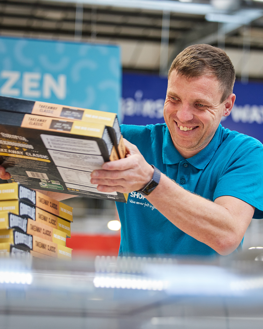 A male Company Shop employee is smiling whilst scanning surplus frozen pizzas through a till.