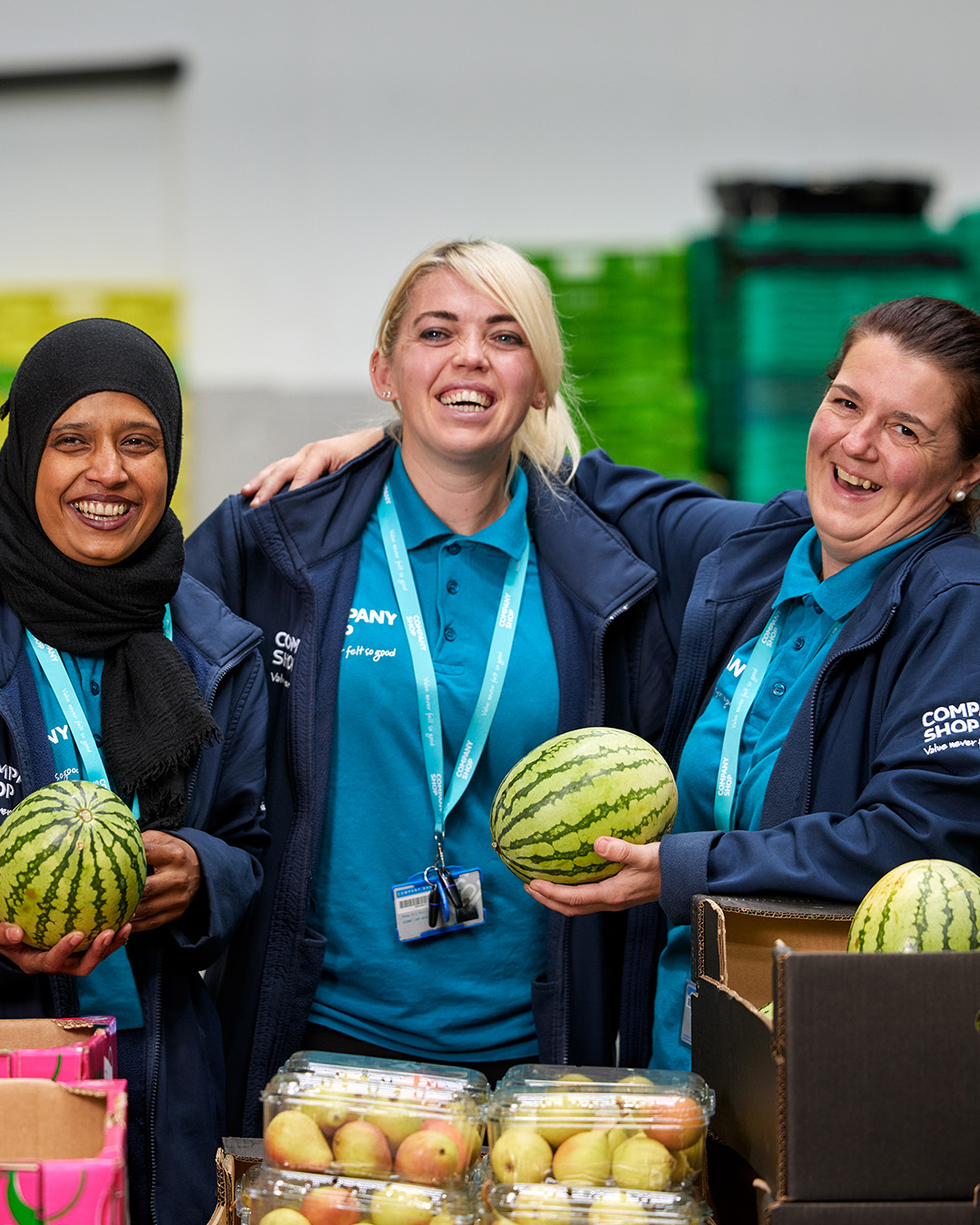 Three female Company Shop employees stand with their arms around each other in front of pallets of fresh fruit.