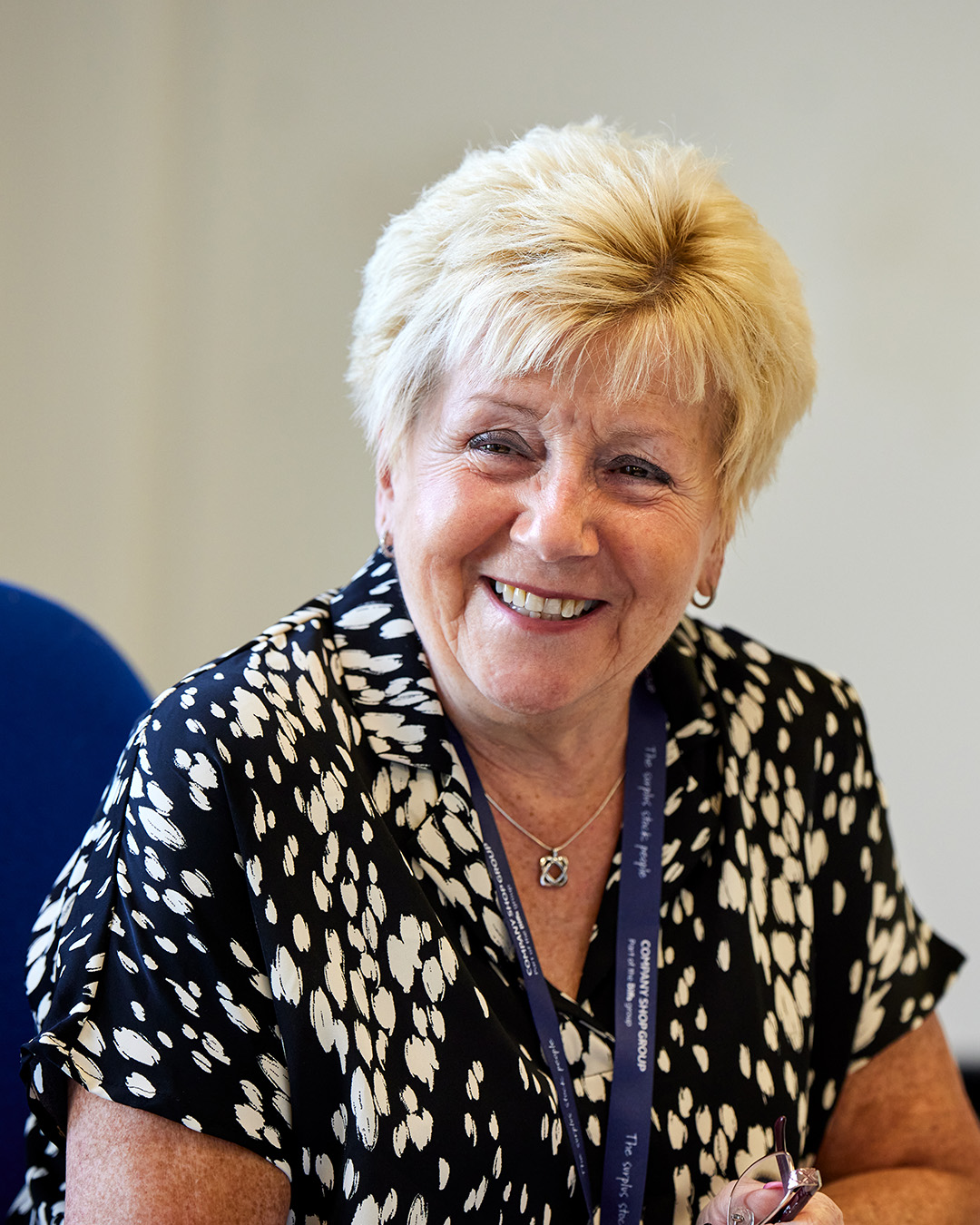 A smiling elderly woman wearing a Company Shop Group lanyard in an office.