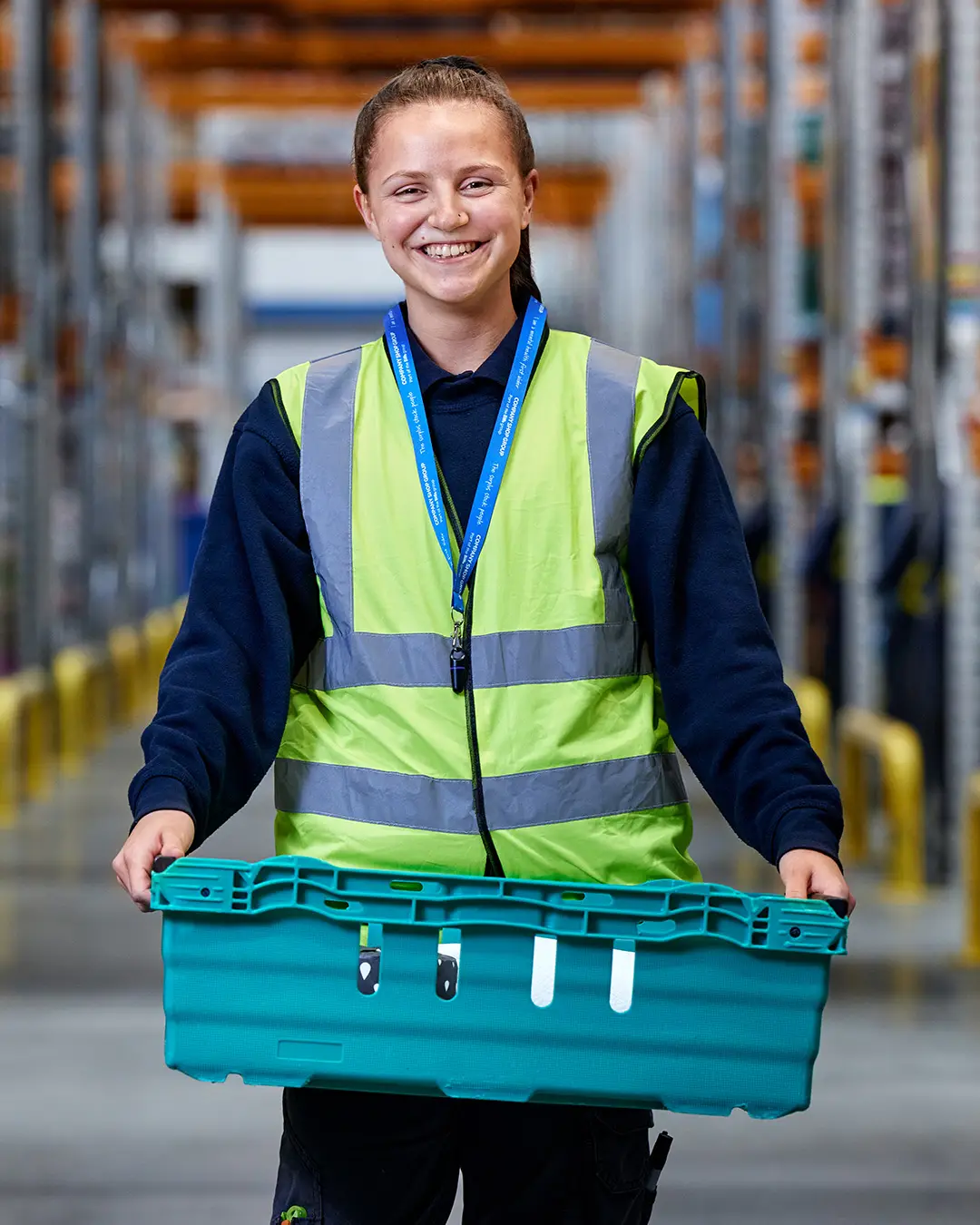 A smiling young woman with a ponytail wearing a yellow high-vis jacket, carrying a green crate in a surplus stock warehouse.