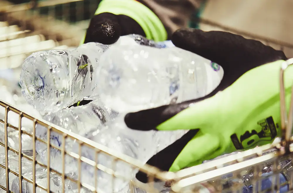 An operative wearing black and yellow gloves is lifting ice-cold plastic bottles of water out of a metal basket.