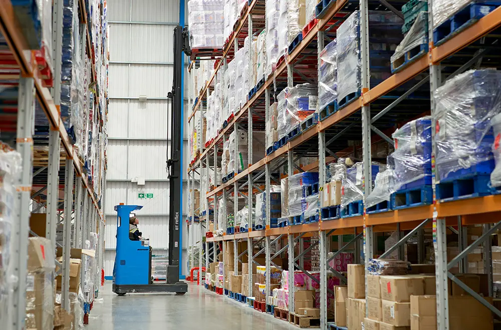 Warehouse operative using a blue forklift to lift a stacked pallet of goods of a high shelf in a warehouse.