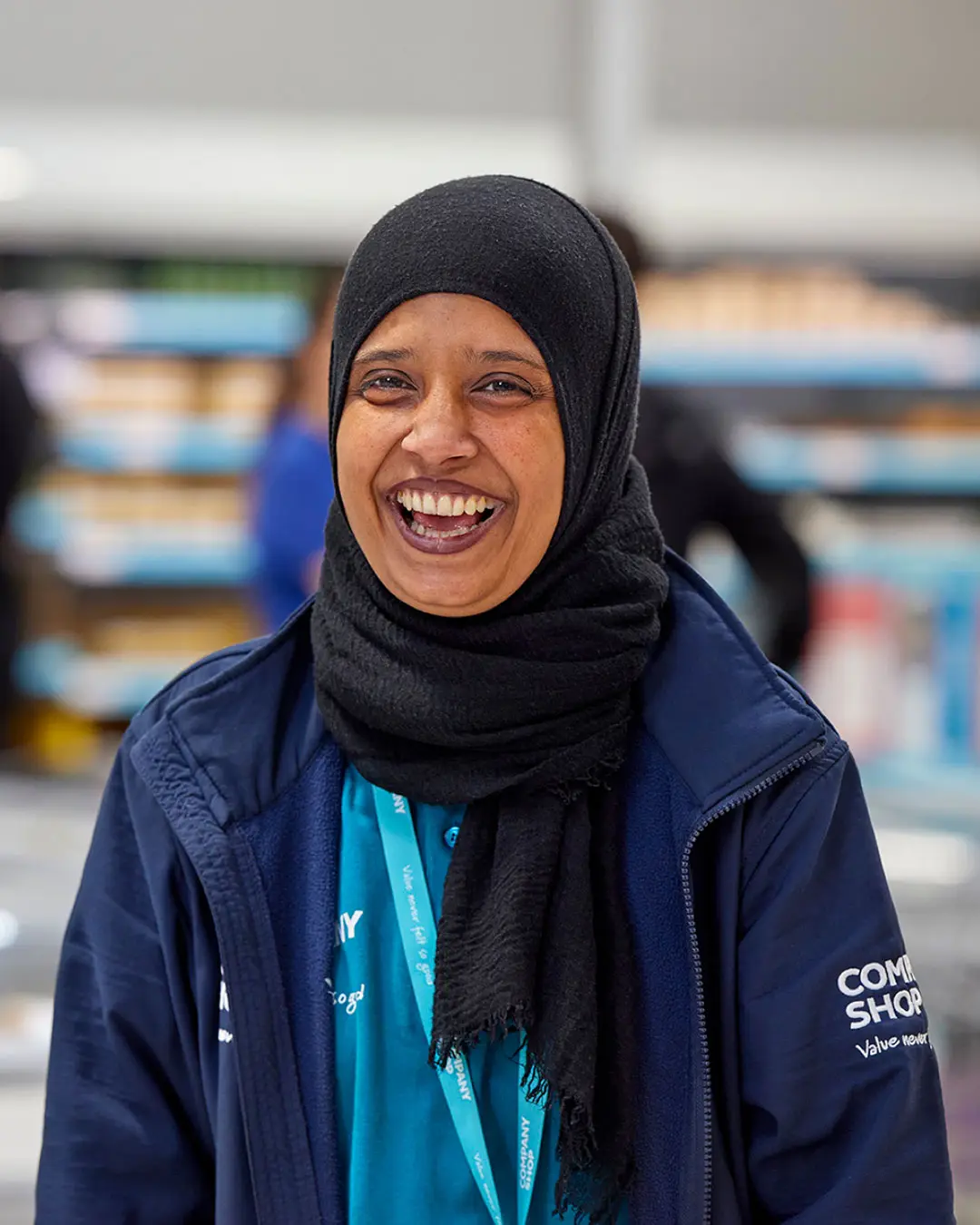 A female Company Shop employee wearing a black hijab and a blue Company Shop jacket smiles widely.
