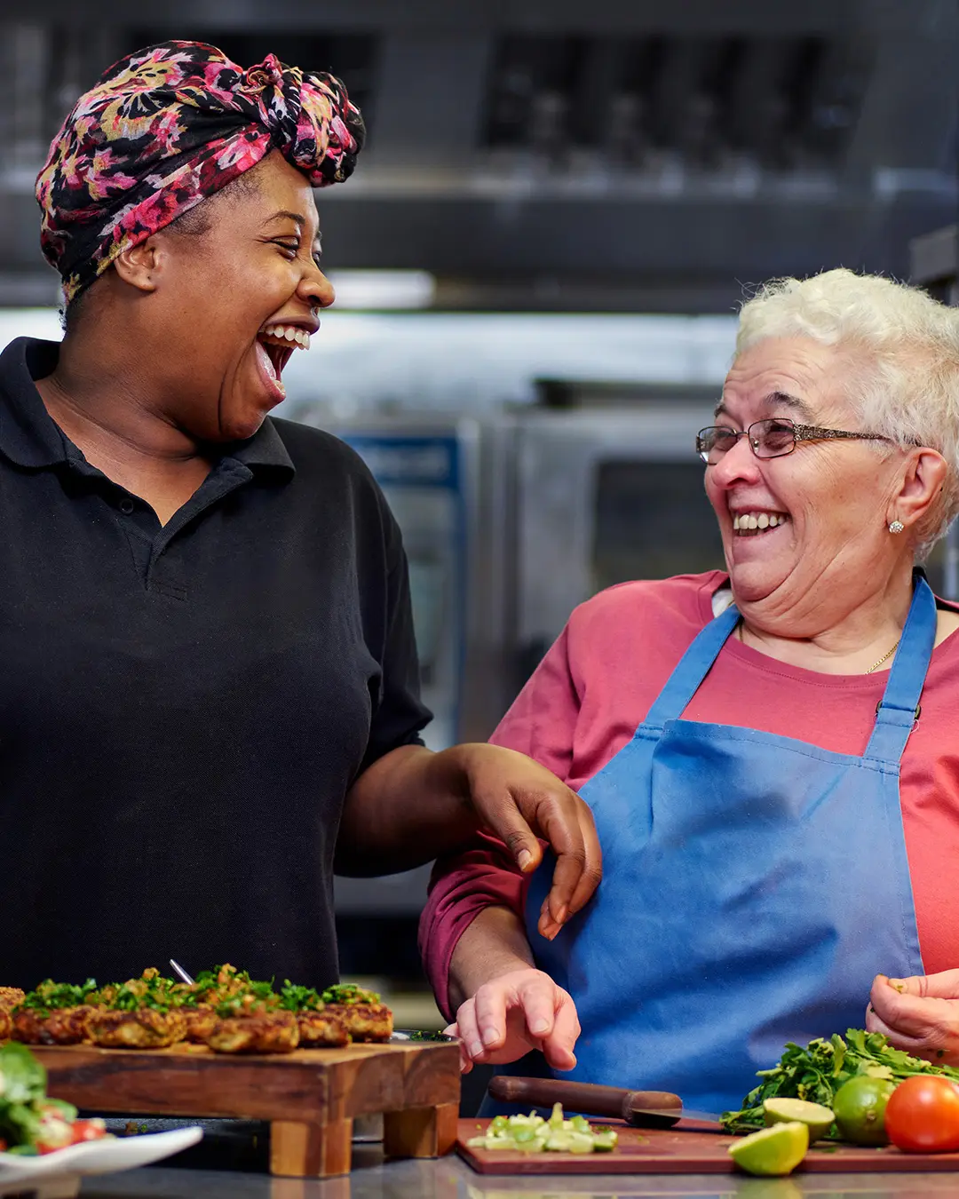 A woman wearing a black t-shirt and a colourful pink head scarf laughs with another woman wearing a blue apron as they prepare tomatoes, avocados and limes at Community Kitchen.