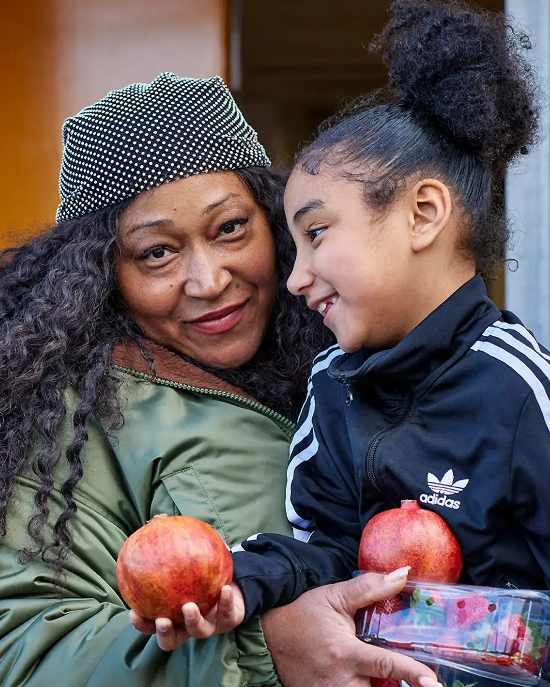 A mother with dark curly hair smiles with her daughter, who is wearing an Adidas jacket outside a Community Shop.