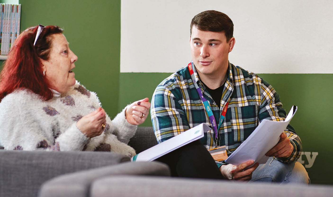 A woman in a white jumper talks animatedly to a man wearing a chequered shirt who looks at her in understanding whilst holding a piece of paper.
