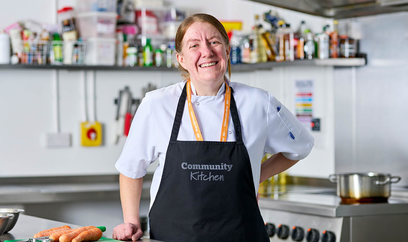 A female chef with red hair, wearing a white Community Kitchen apron, smiles as she prepares carrots.