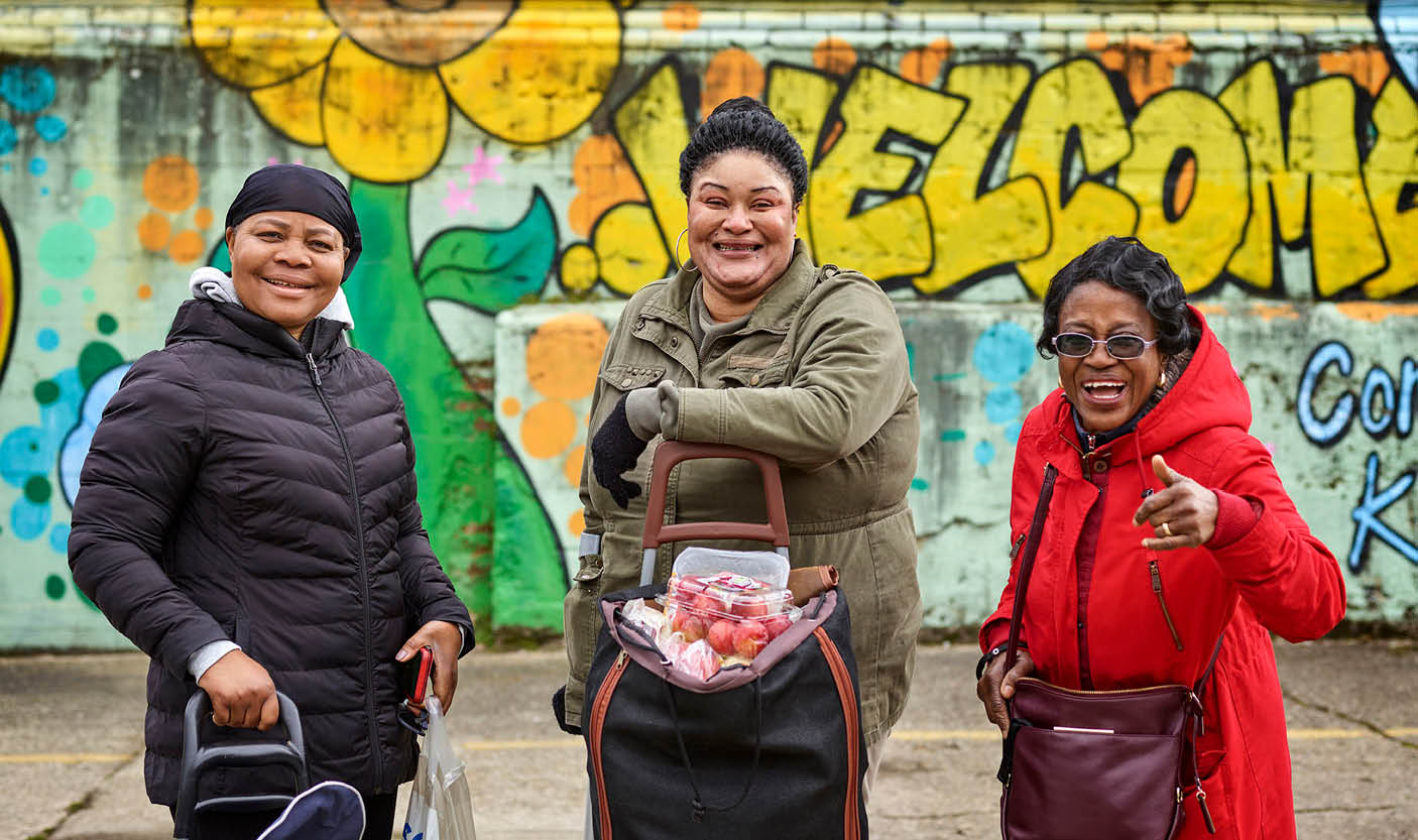 Three women with 'granny shoppers' filled with fresh food smile in front of a wall decorated with colourful graffiti.