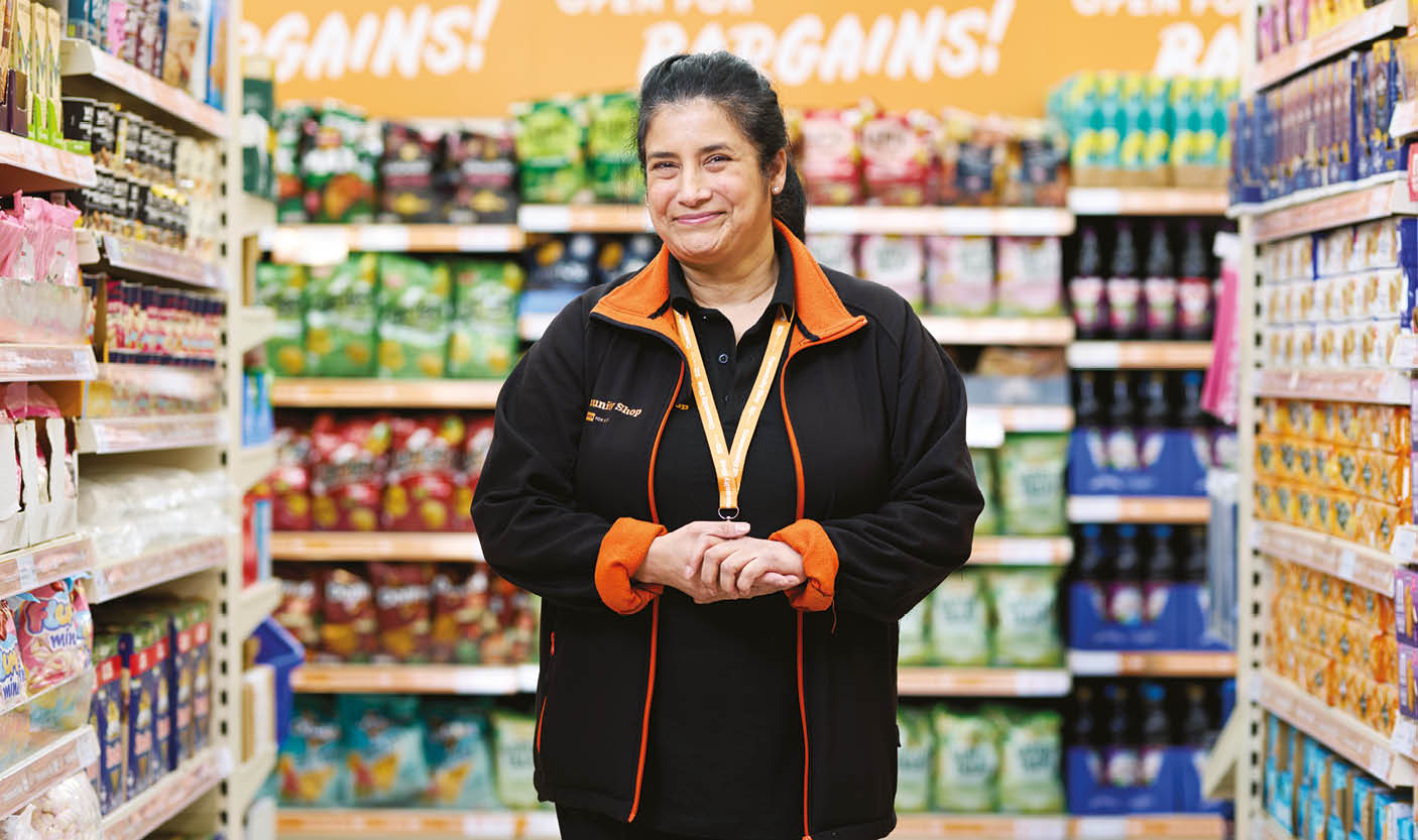 An asian woman in a black Community Shop uniform smiles inside a community store.