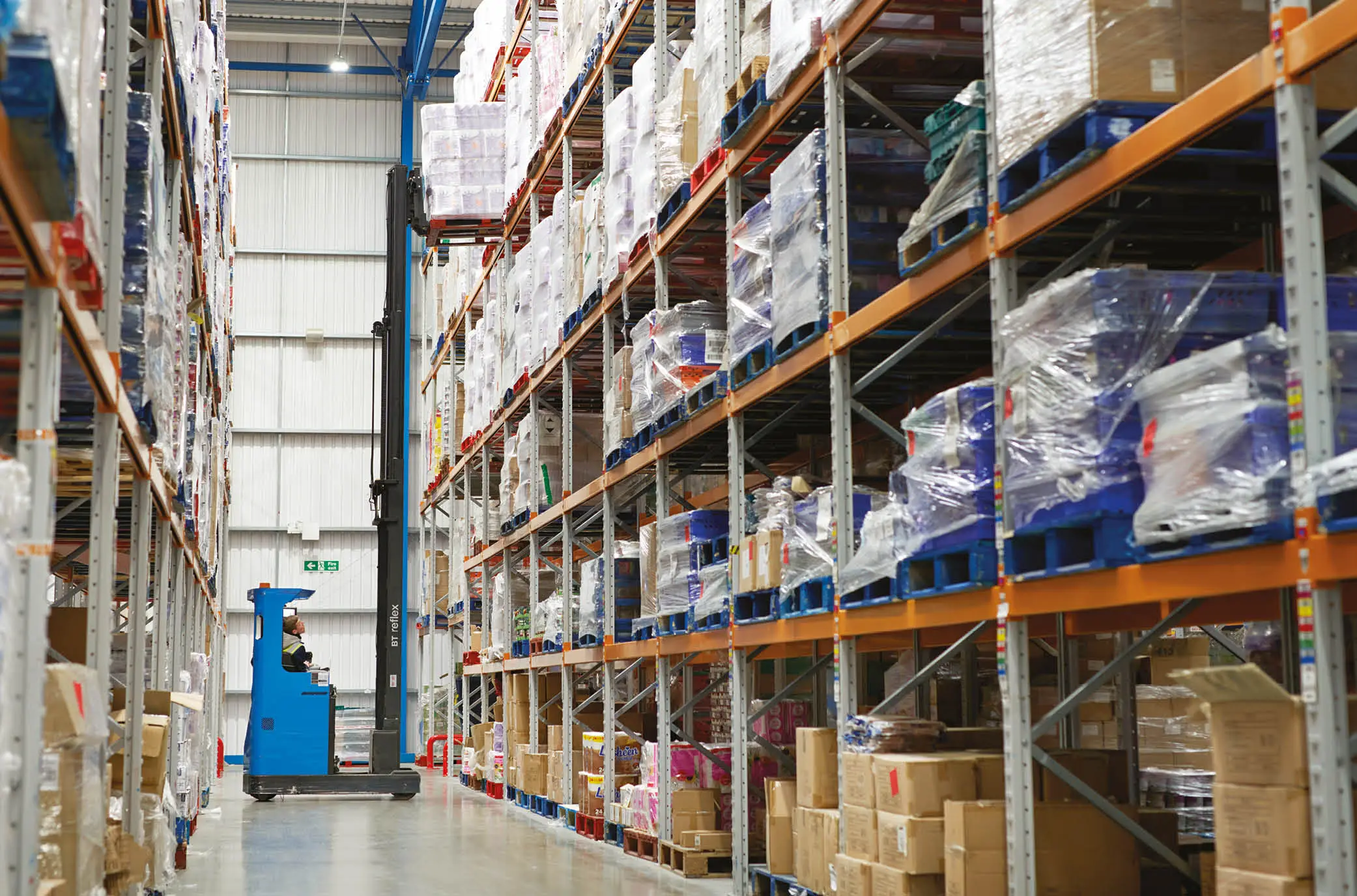 Warehouse operative using a blue forklift to lift a stacked pallet of goods of a high shelf in a surplus warehouse.