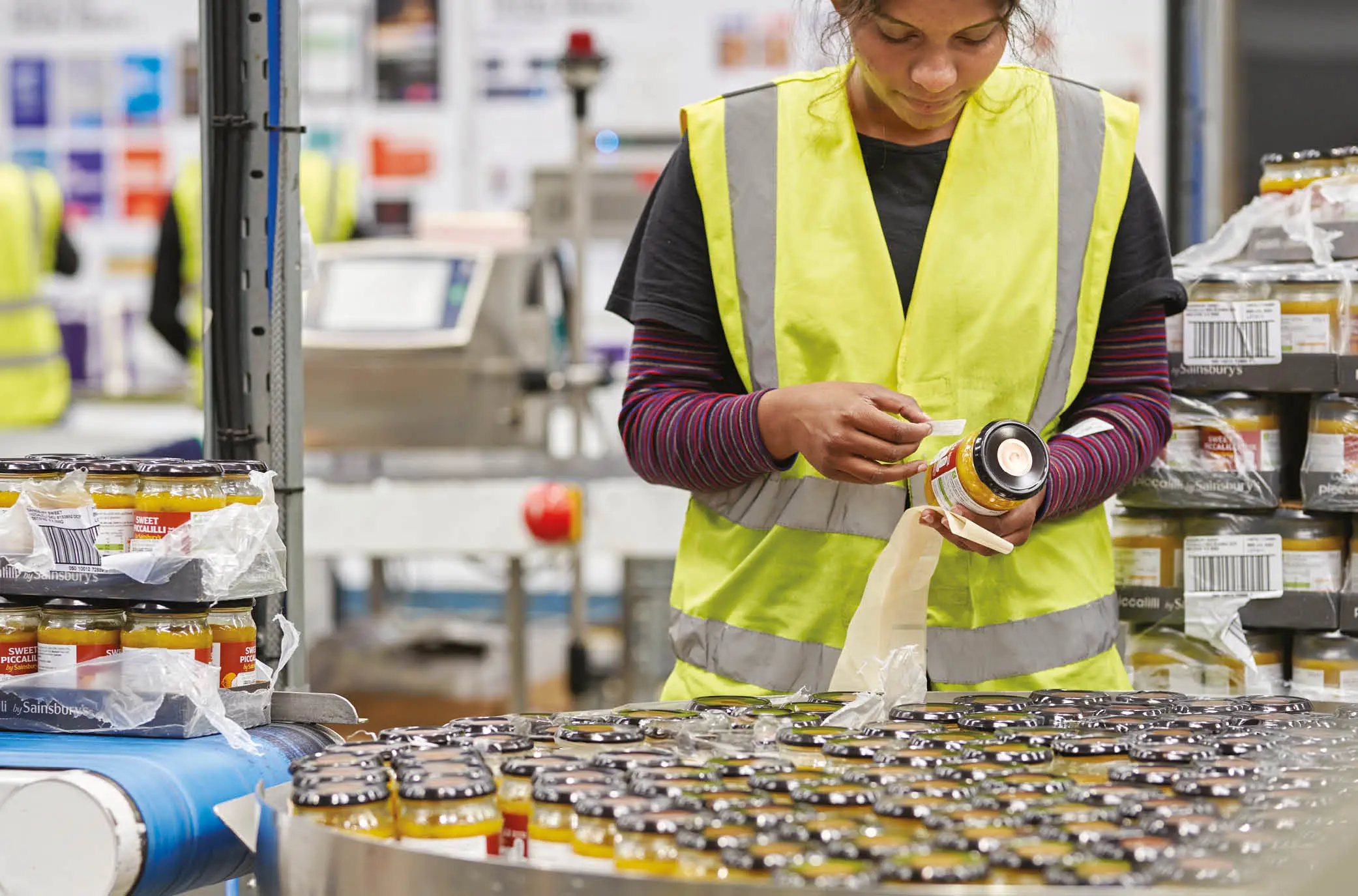 Female warehouse operative wearing a yellow high-vis jacket, carefully re-labelling jars of apple sauce.