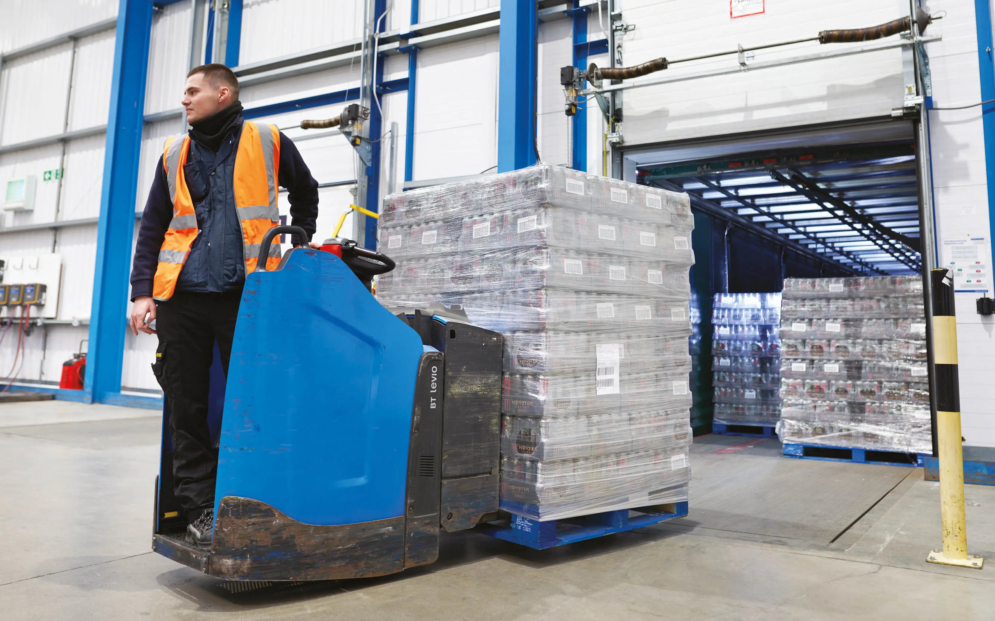 Company Shop Group warehouse operative wearing an orange high-vis jacket powering a forklift loaded with a pallet of energy drinks.