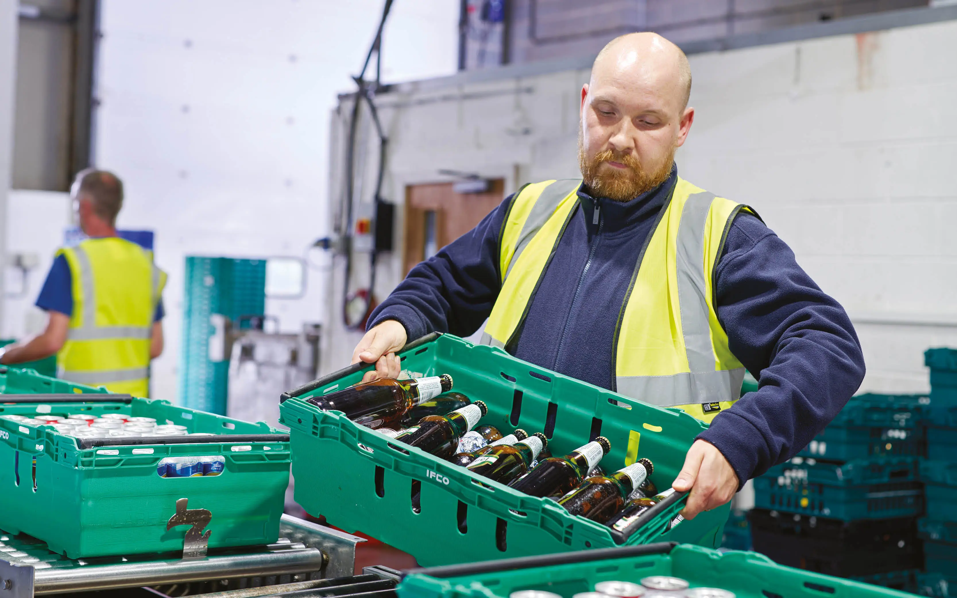 Man wearing a yellow high-vis jacket giving full concentration to the crate of glass beer bottles he is carrying.