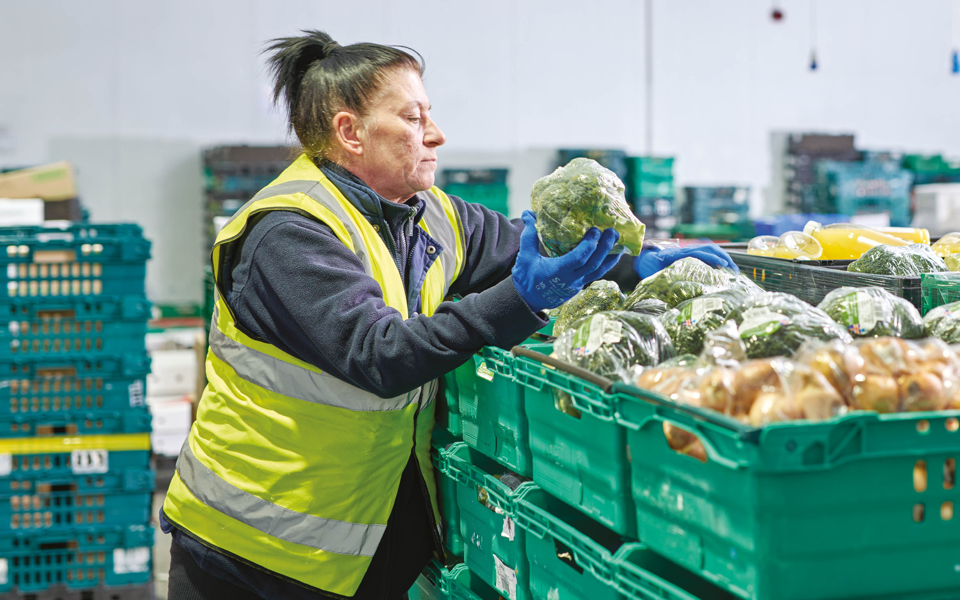 A female warehouse operative in a yellow high-vis jacket is loading green crates with surplus broccoli.