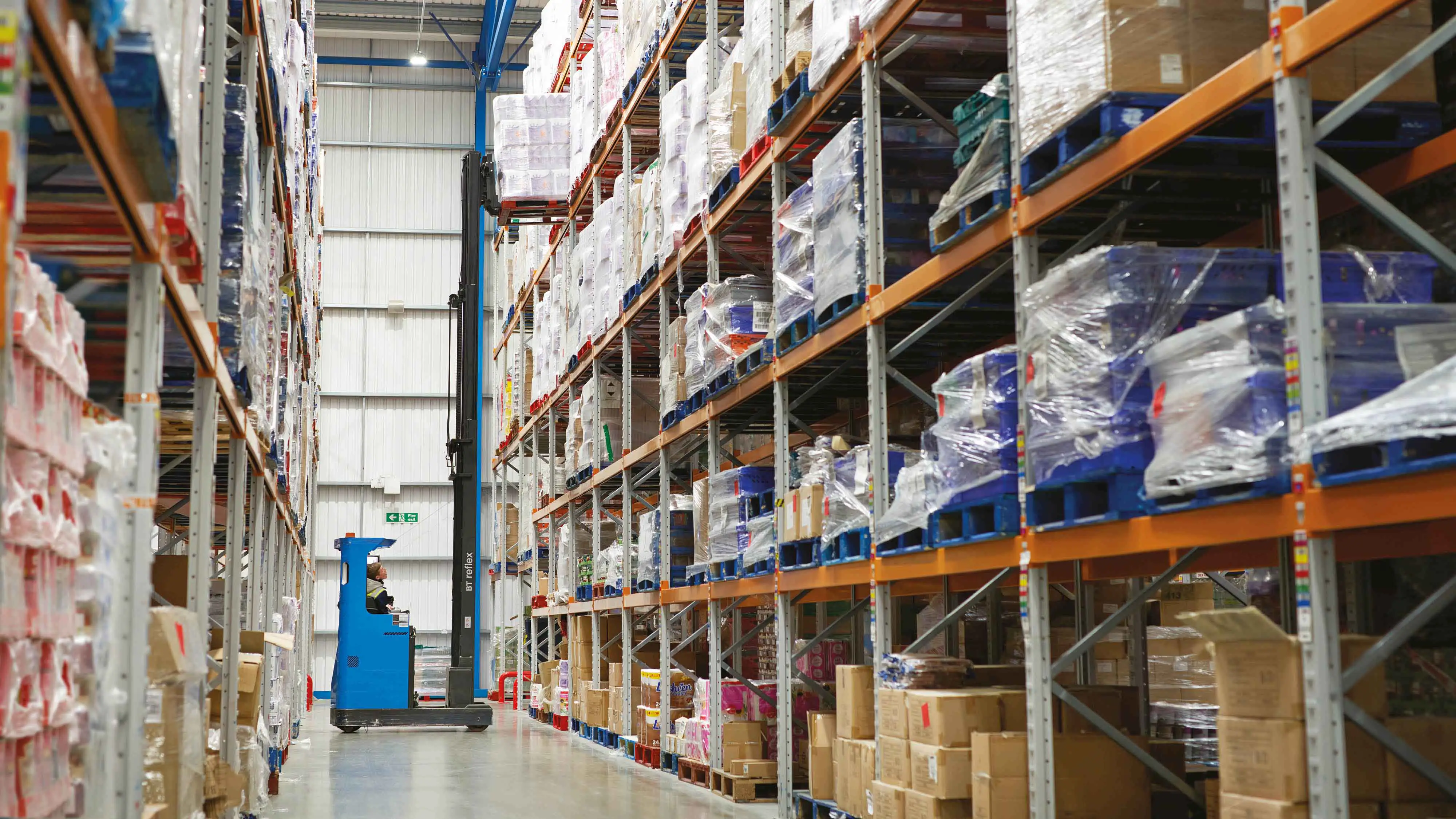 Warehouse operative using a blue forklift to lift a stacked pallet of goods on a high shelf in a surplus warehouse.